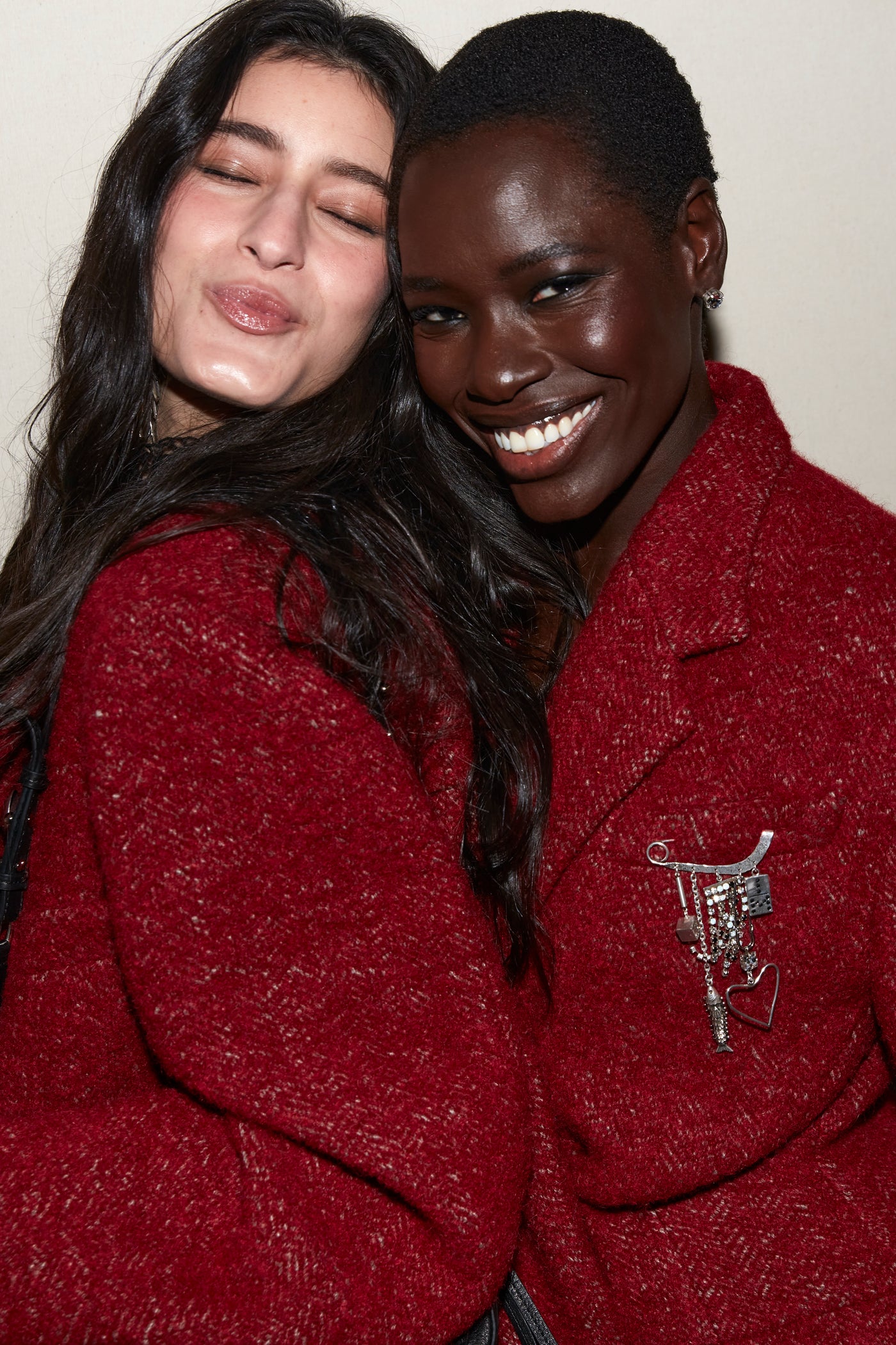 Models both wear deep red coats styled with a Brooch with safety pin pendants with stones, hearts, and chains