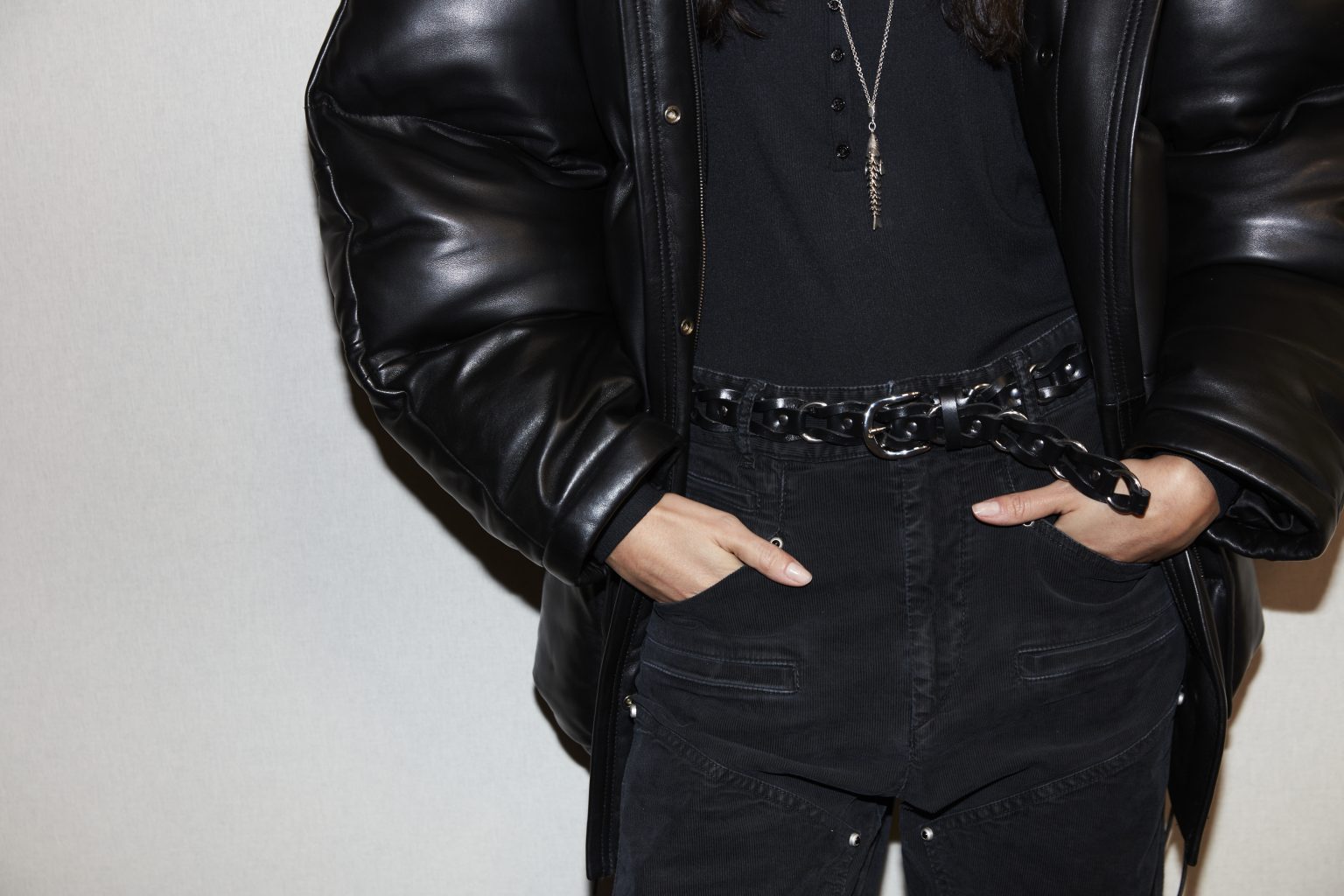 Female midsection with hands in pockets, wearing a shiny black puffy jacket, t-shirt and washed jeans with a braided belt, all in black.