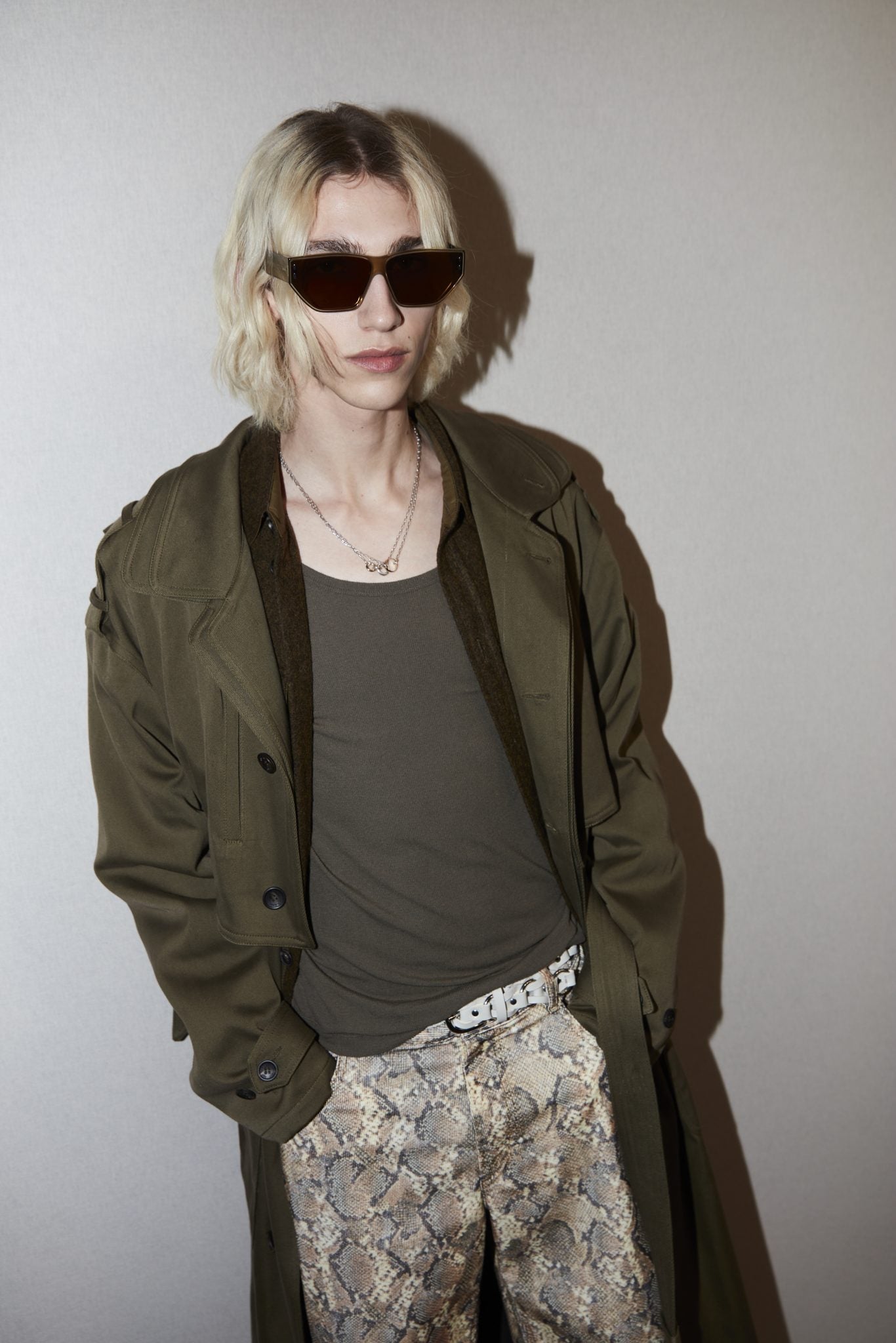 Cropped backstage image of a model wearing sunglasses, a khaki tee and trench, black and tan snakeskin-printed jeans and a white studded belt.