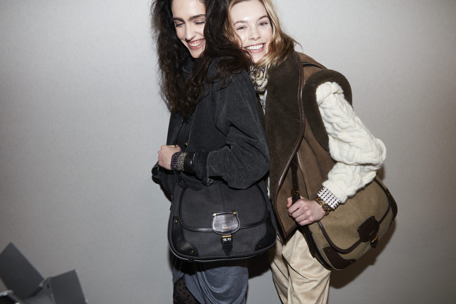 Backstage image of two female models, one wearing shades of black and grey, the other in tan and brown, both with messenger bags.