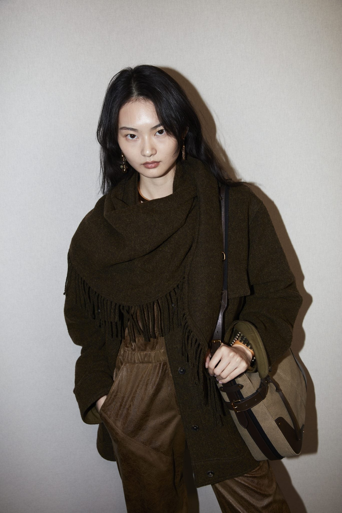 Female model wearing a dark brown jacket and fringed scarf over a chocolate brown jumpsuit and carrying an oversized tan messenger bag.