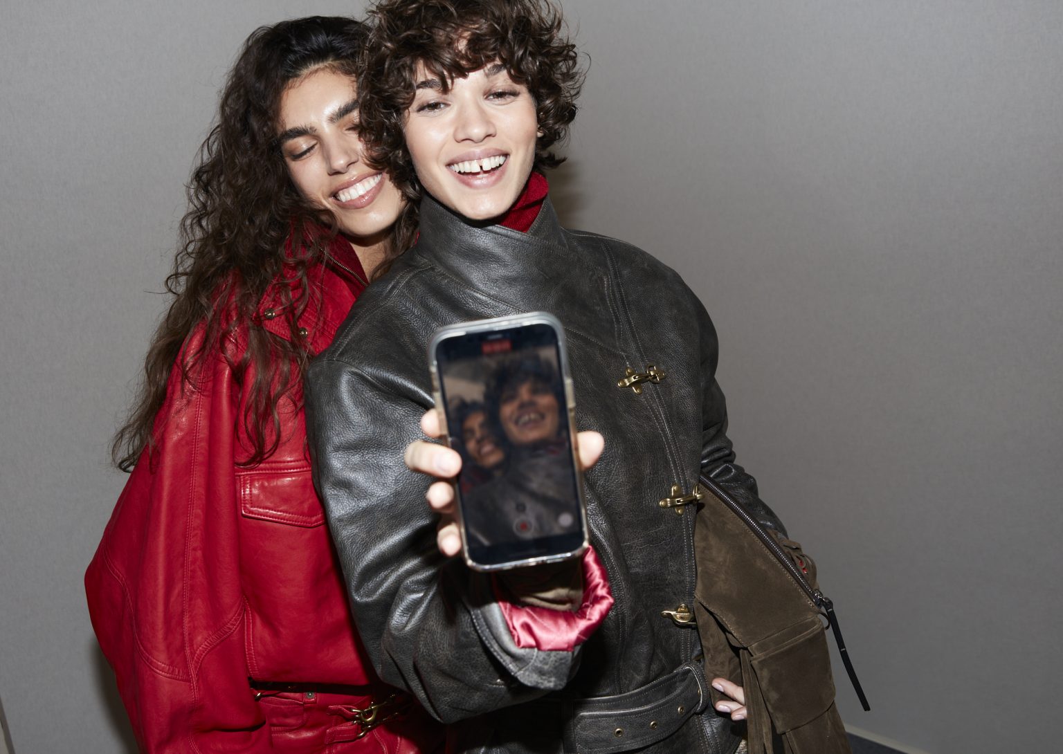 Two models smiling, one wearing a black leather jacket with clip fasteners, flashing a just-taken selfie. The other wears a red jacket. 