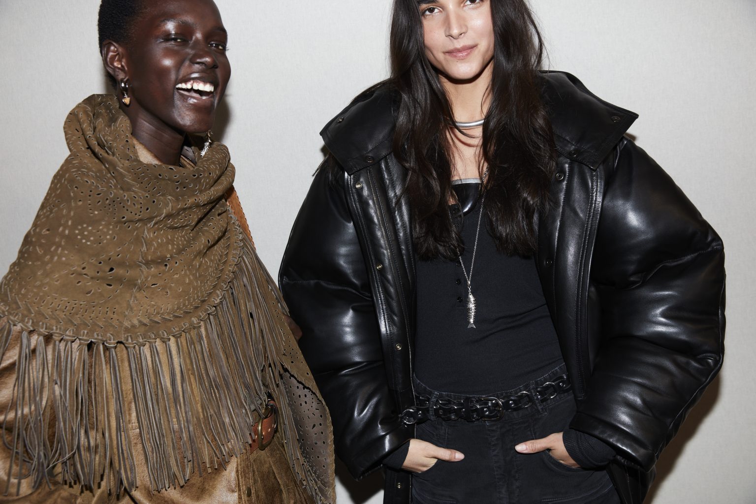 Cropped image of two models backstage, one in a brown fringed scarf and brown jacket, the other in a puffy black jacket.