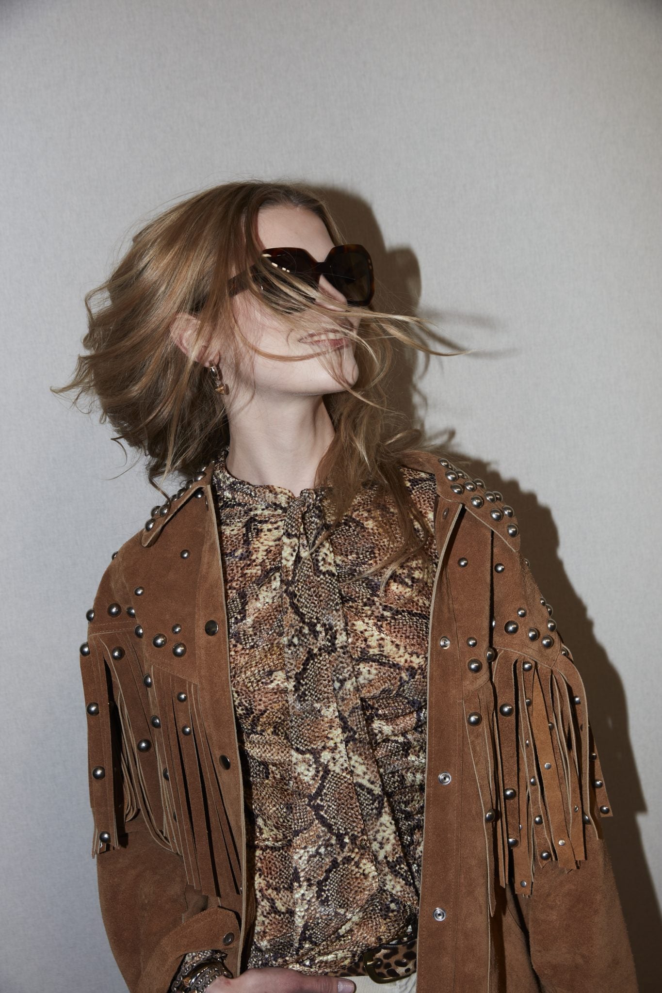 Cropped backstage image of female model in large sunglasses, a light brown suede fringed jacket with metallic bead details and a snakeskin-printed blouse.
