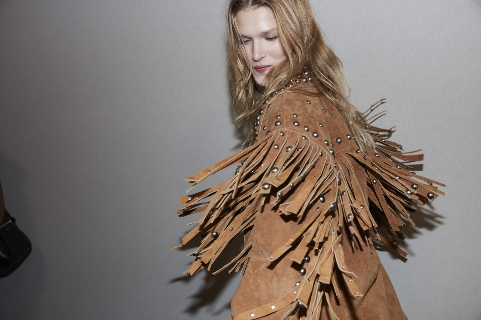 Cropped backstage image of female model wearing a light brown suede fringed jacket with metallic bead details, the fringes flying as she spins.