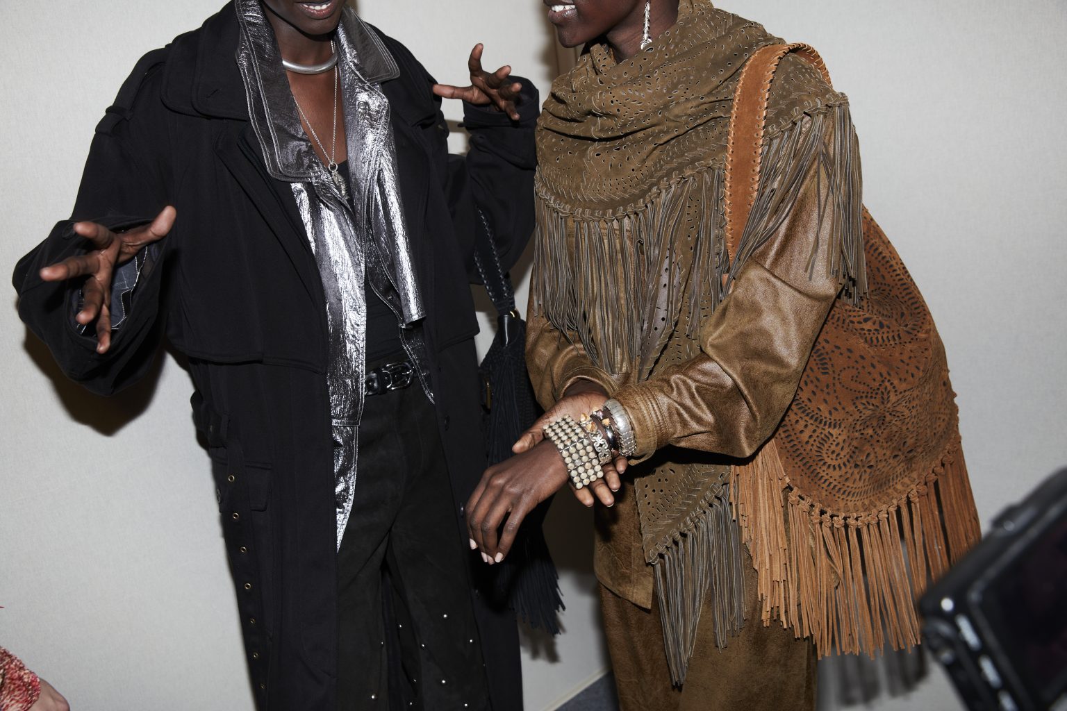 Backstage image of two models’ midsections, one in brown clothing with various fringe details, one in a silver blouse under a black jacket.
