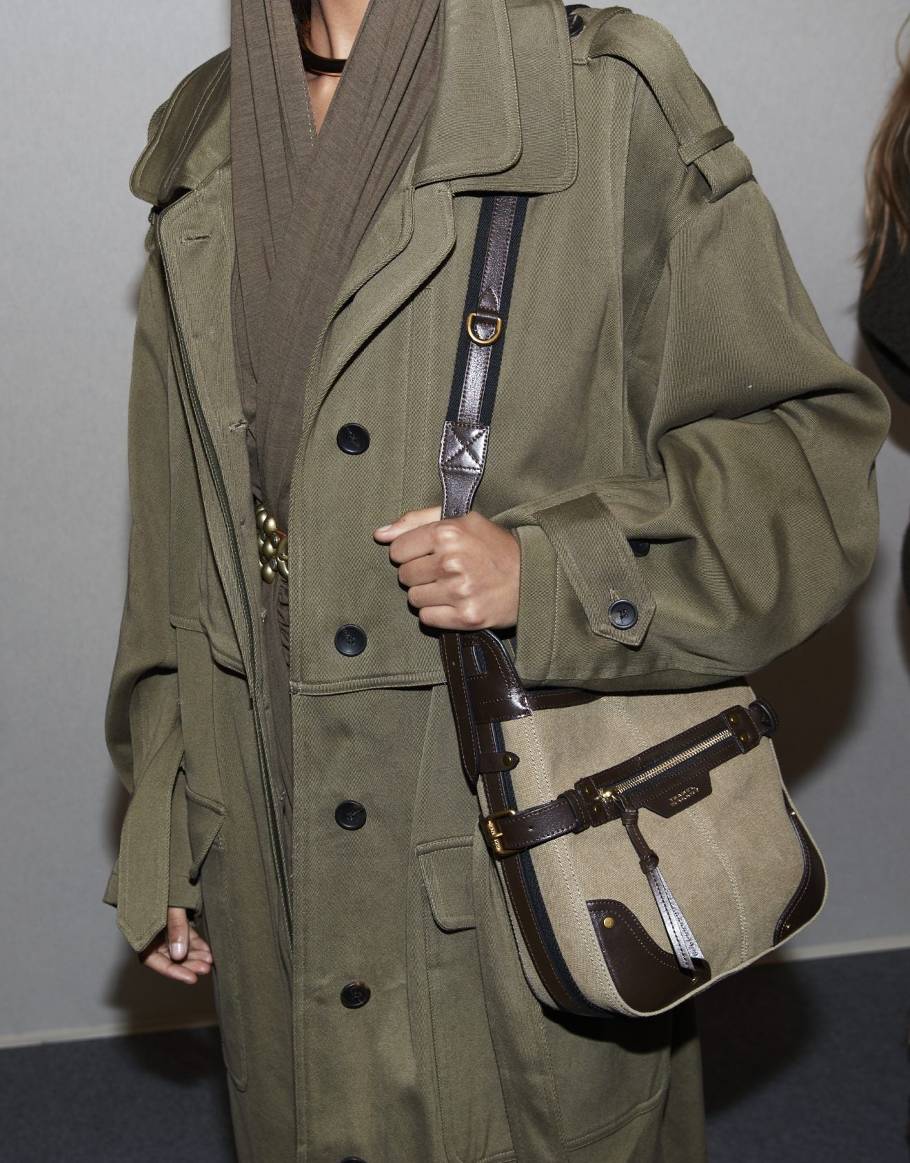 Backstage image of a model’s midsection dressed in an oversized khaki trench and carrying a tan messenger bag with dark brown leather trim. 
