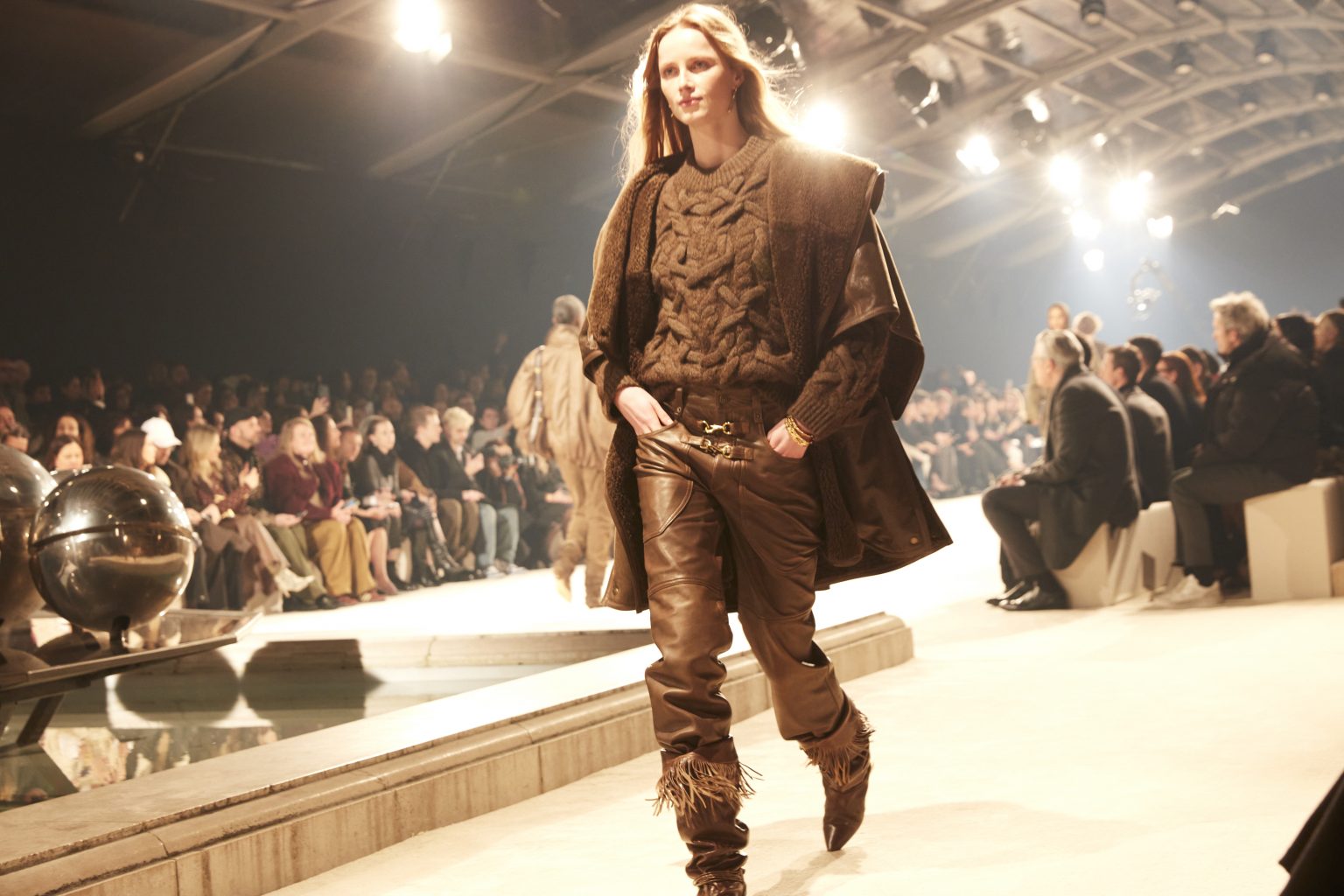 Female model on catwalk wearing a chunky cable-knit sweater, oversized vest, leather pants and fringed boots, all in chocolate brown.