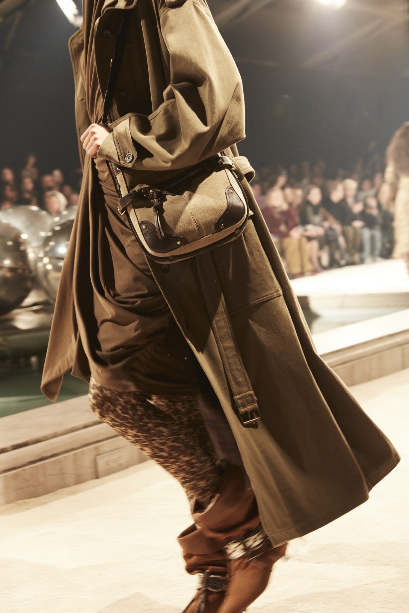 Cropped catwalk image of model wearing an oversized tan trench, brown gathered dress, leopard-printed tights and slouchy brown boots, carrying a messenger bag.