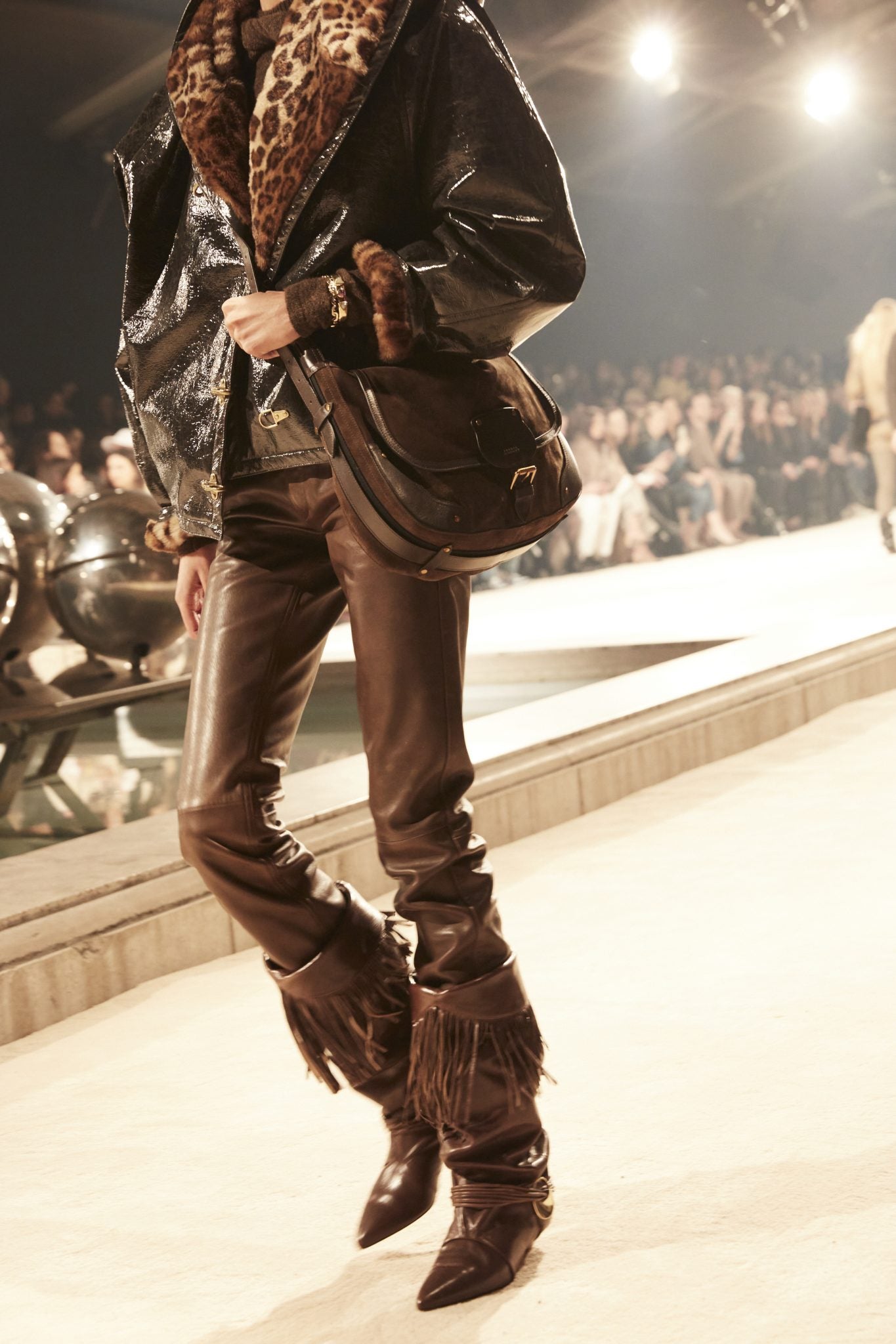 Cropped image of female model wearing a black faux leopard fur-lined coat and brown leather pants, tucked into brown fringed leather boots.