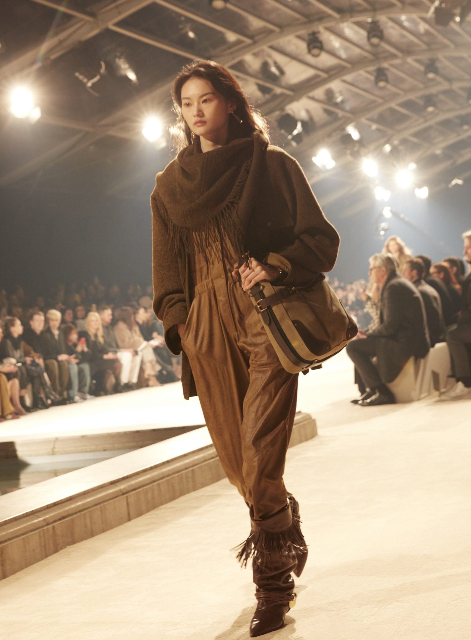 Female model on catwalk wearing a jumpsuit with waist gathers, fringed scarf, sweater and fringed boots, carrying a large bag, all in brown.