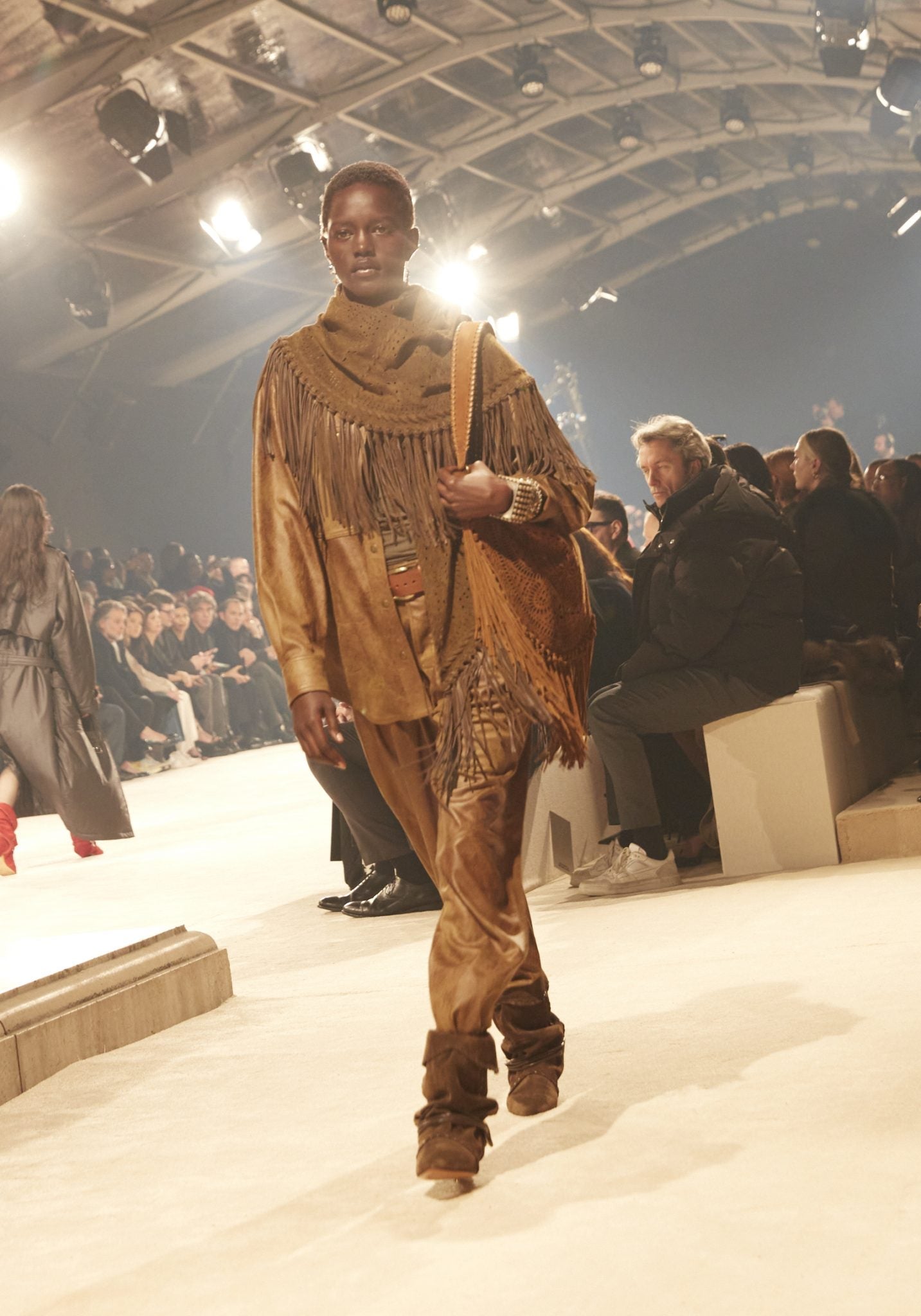 Female model on catwalk wearing a fringed scarf, overshirt and pants tucked into boots, carrying a large fringed bag, all in various browns.