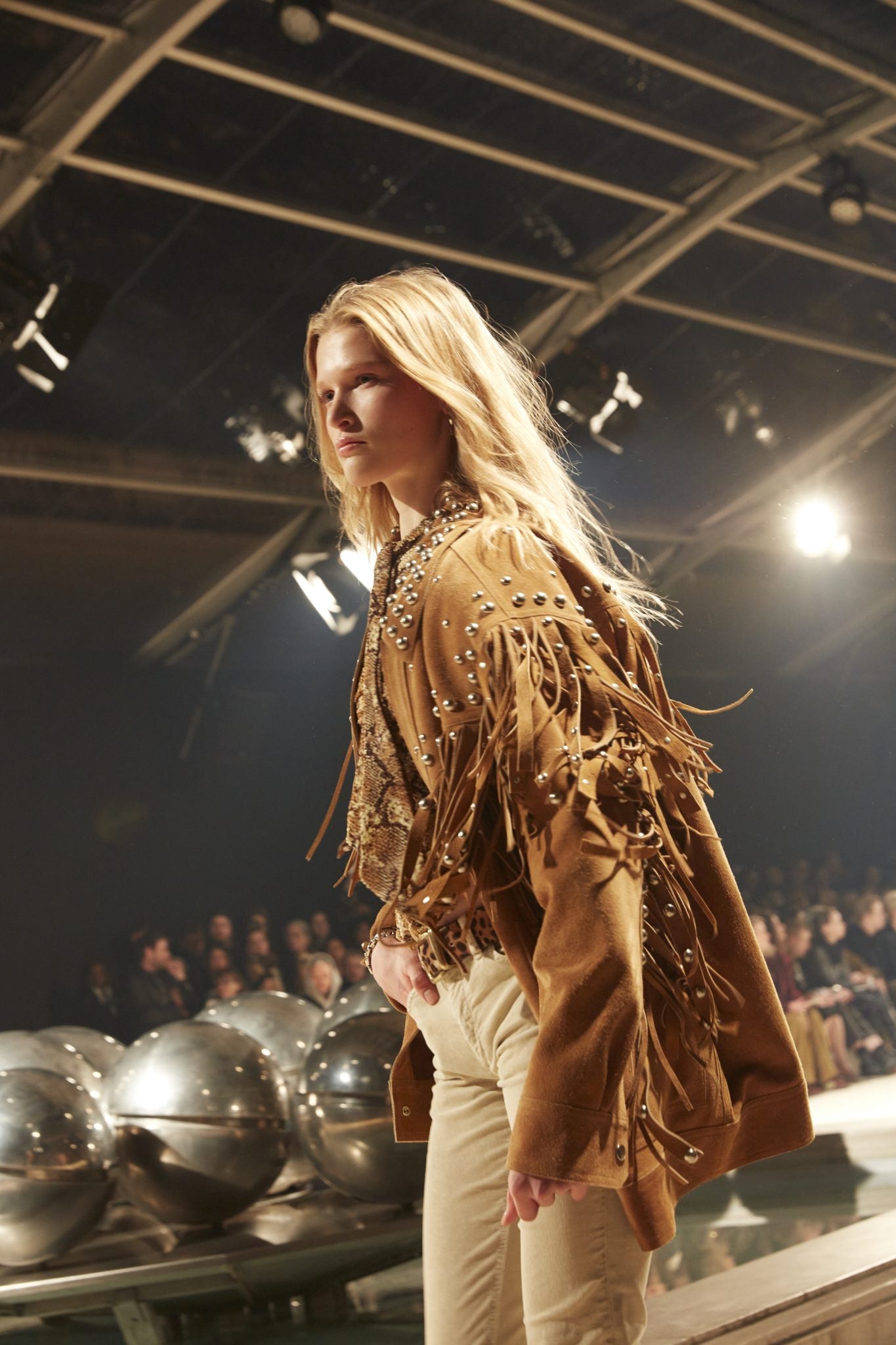 Cropped image of female model on catwalk wearing a tan suede fringed jacket with metallic bead details, a leopard-print belt and cream-colored jeans.