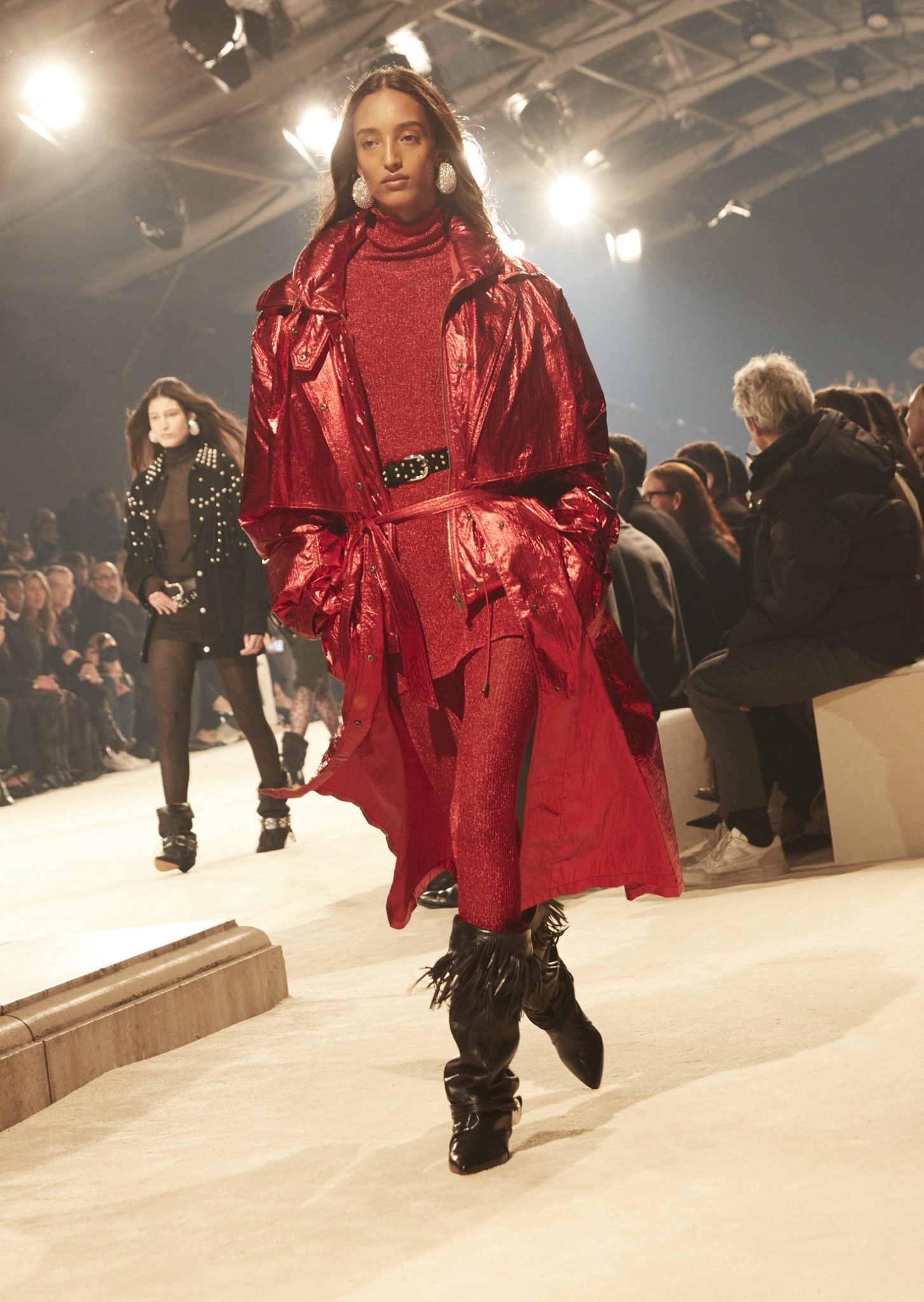 Female model on catwalk wearing an iridescent red trench, a red turtle-neck tunic and tights, studded black belt and black fringed boots.