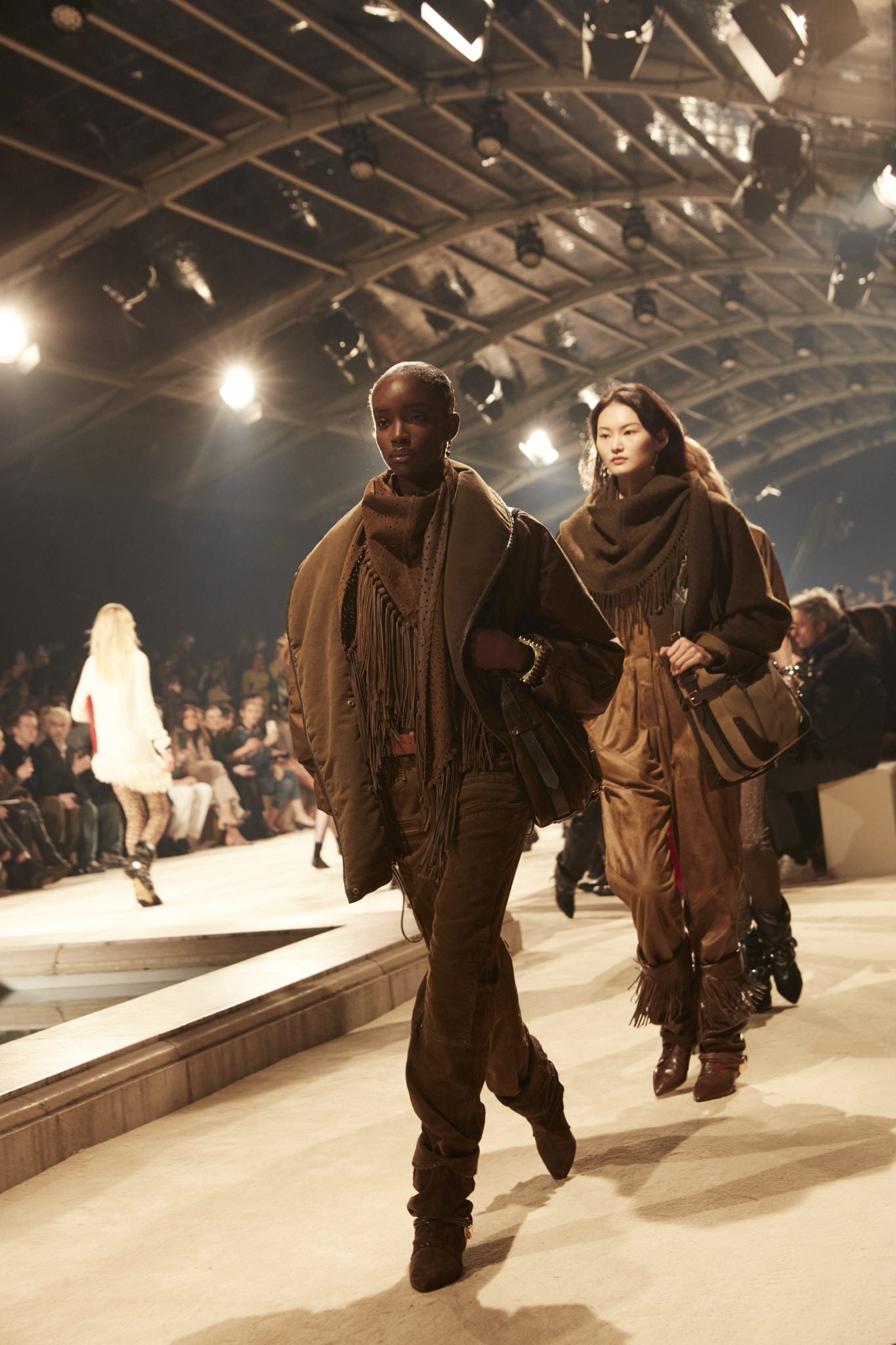 Two female models on catwalk dressed in shades of brown, both wearing fringed scarves, carrying messenger bags and wearing brown boots.