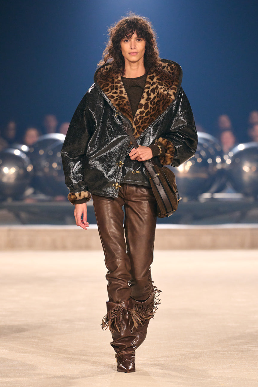 Female model on catwalk wearing a black coat lined in faux leopard with chocolate brown leather pants tucked into brown fringed boots.