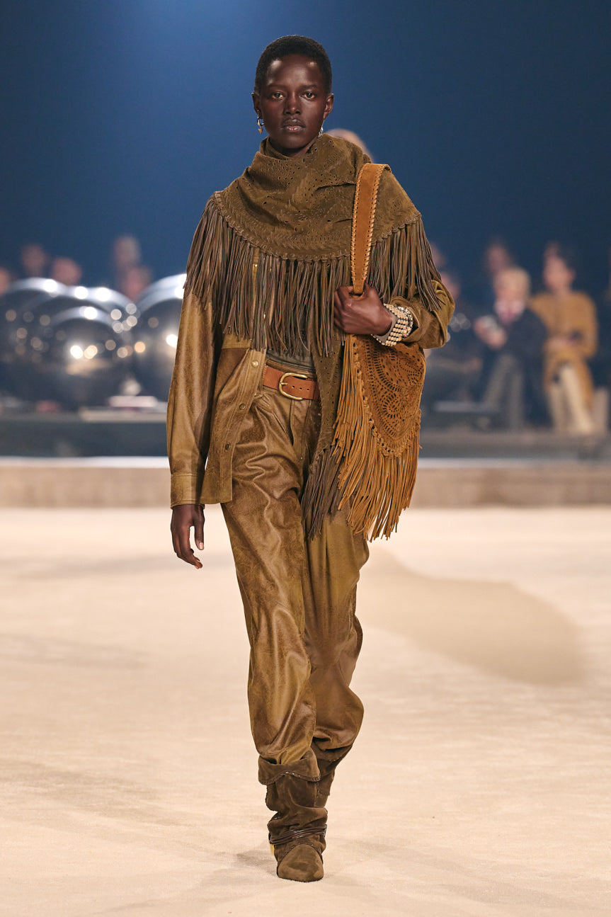 Image of female model on catwalk wearing a tan overshirt and trousers with a brown suede fringed scarf, carrying a tan fringed bag.