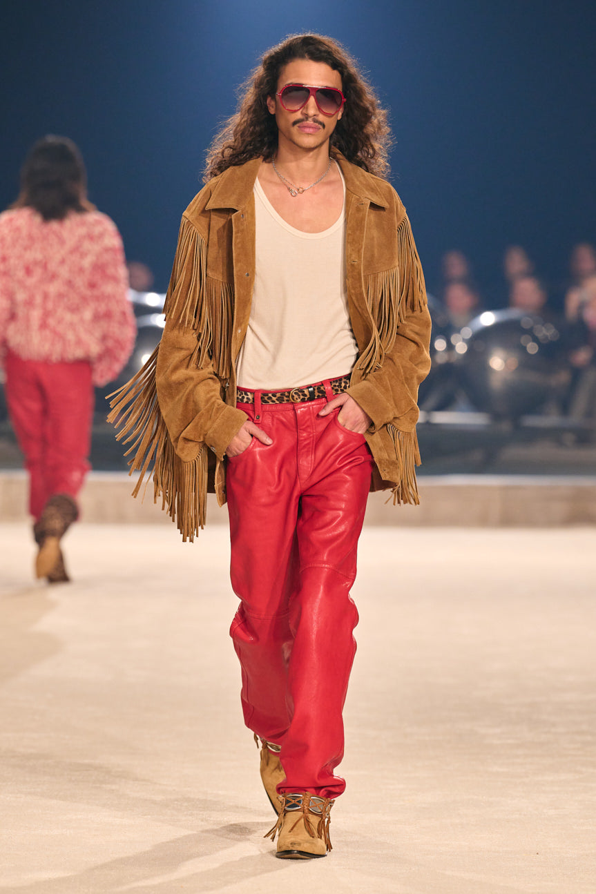 Male model on catwalk wearing a cream-colored scoop-neck tee, tan suede fringed jacket, baggy red leather jeans and tan ankle boots.