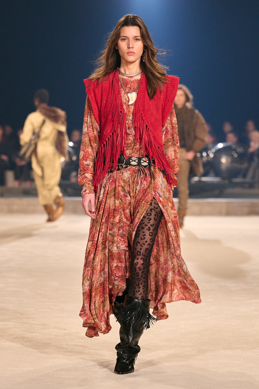 Female model on catwalk wearing a printed red midi dress under a red fringed vest with black leopard tights and slouchy black boots.