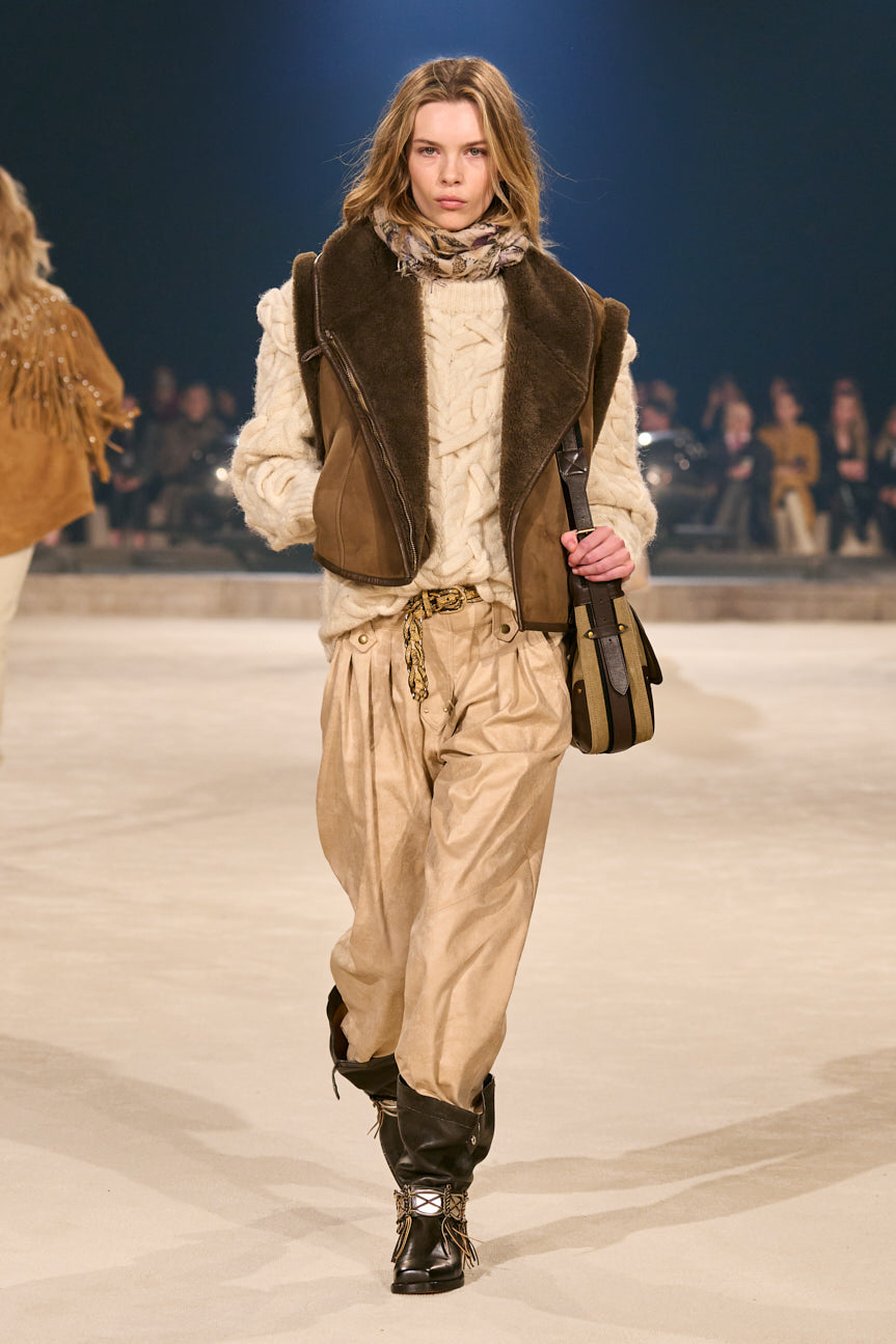 Image of female model on catwalk wearing a tan chunky cable-knit sweater with gathered tan pants tucked into slouchy, dark brown boots.