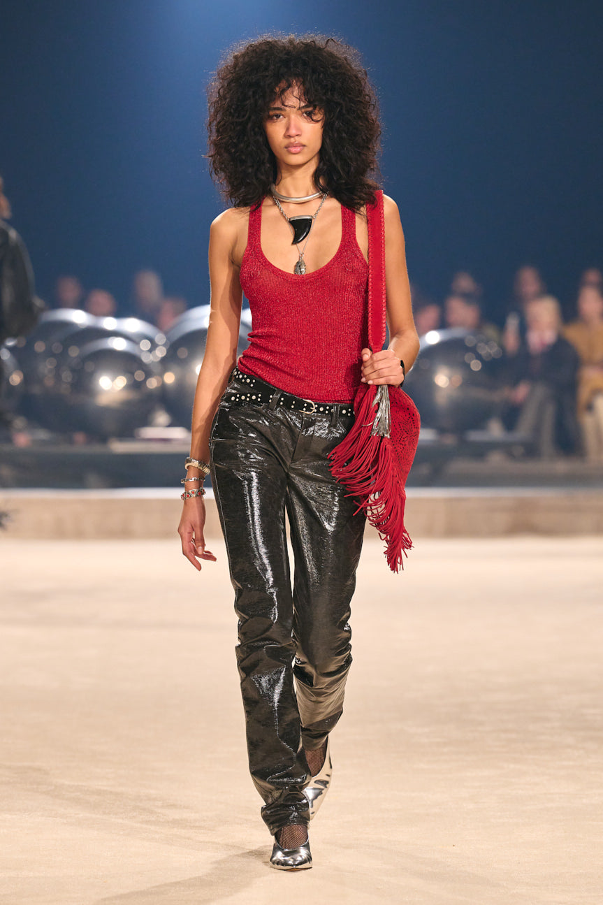 Image of female model on catwalk wearing a red tank, shiny black pants, studded black belt and carrying a red fringed bag. 