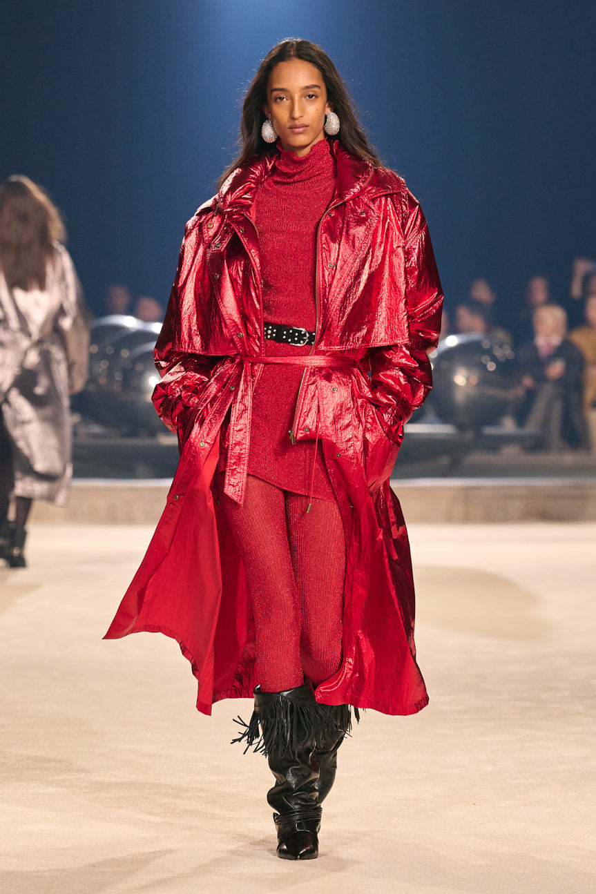 Female model on catwalk wearing an iridescent red trench over a red turtleneck tunic and tights, studded black belt and black fringed boots.