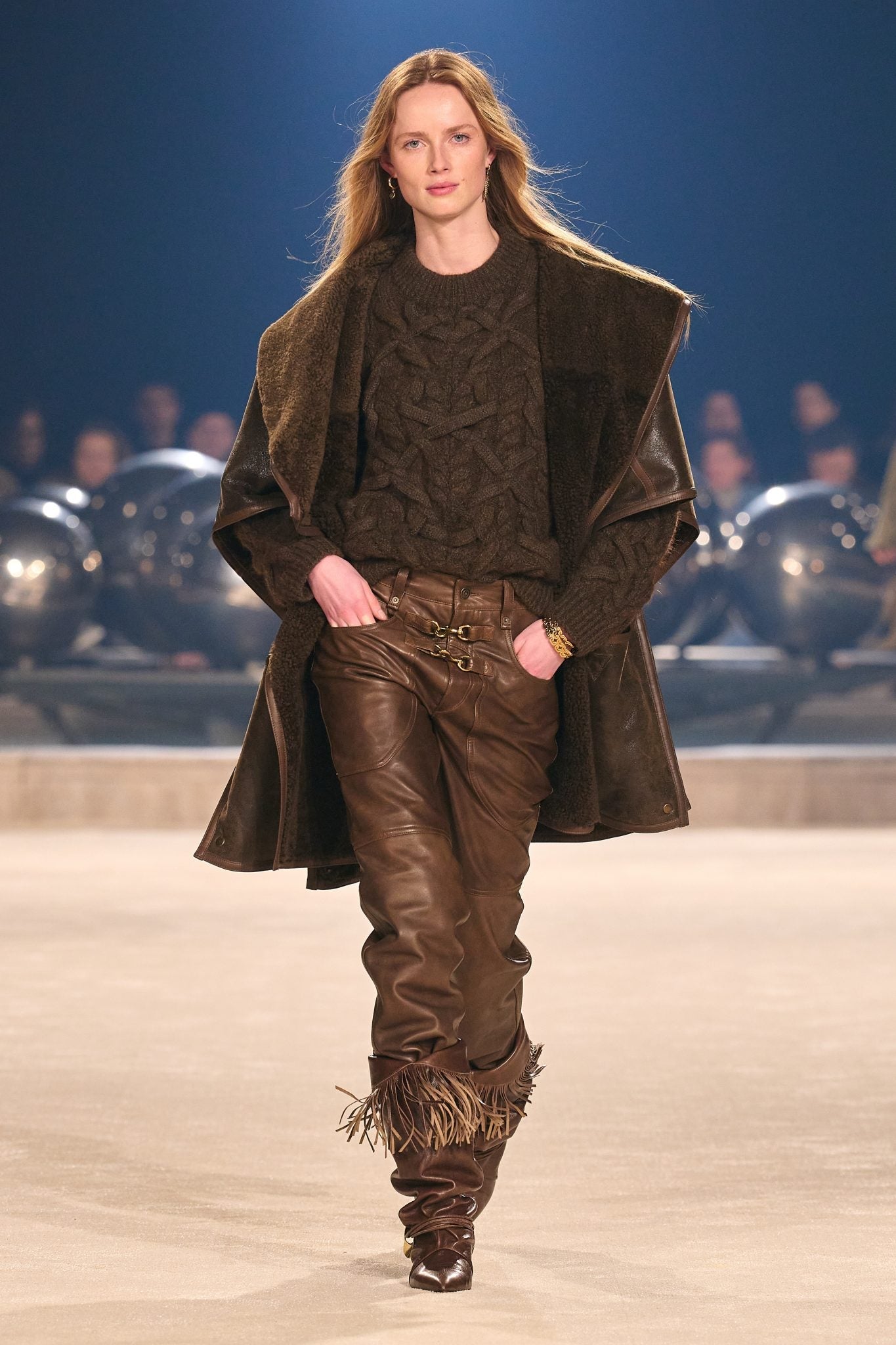 Female model on catwalk wearing a chunky cable-knit sweater under an oversized coat with leather pants tucked into fringed boots, all in brown.