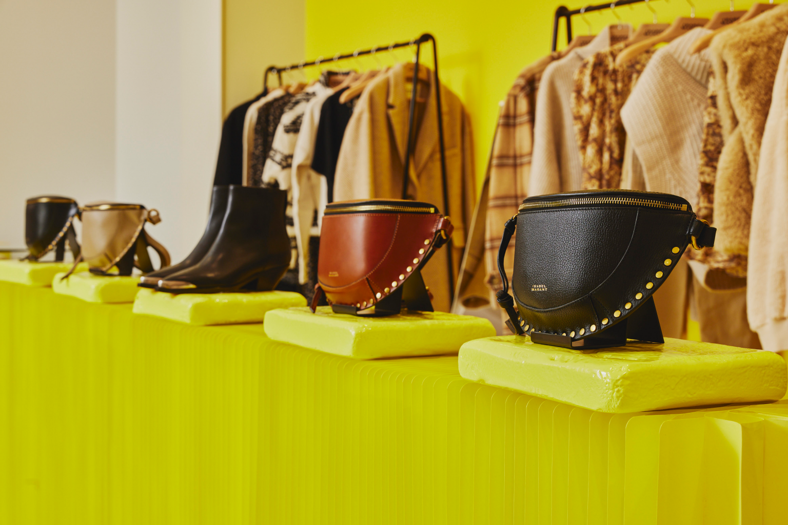 Close-up image of yellow display stands featuring various pieces from the Isabel Marant clothing collection, including handbags and boots, with clothing visible in background.