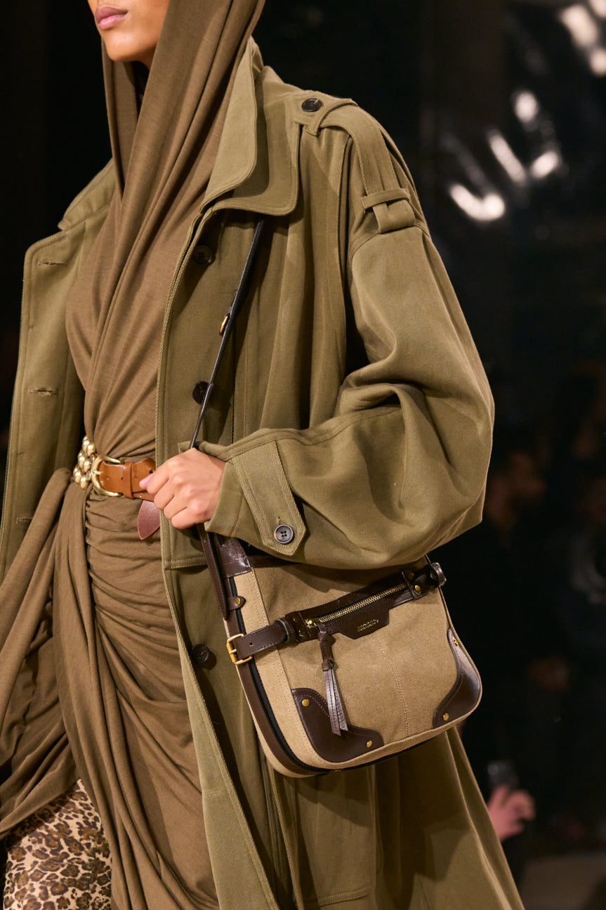 Cropped image of female model wearing a belted oversized hooded tunic under a khaki trench, carrying a tan messenger bag with brown trim.