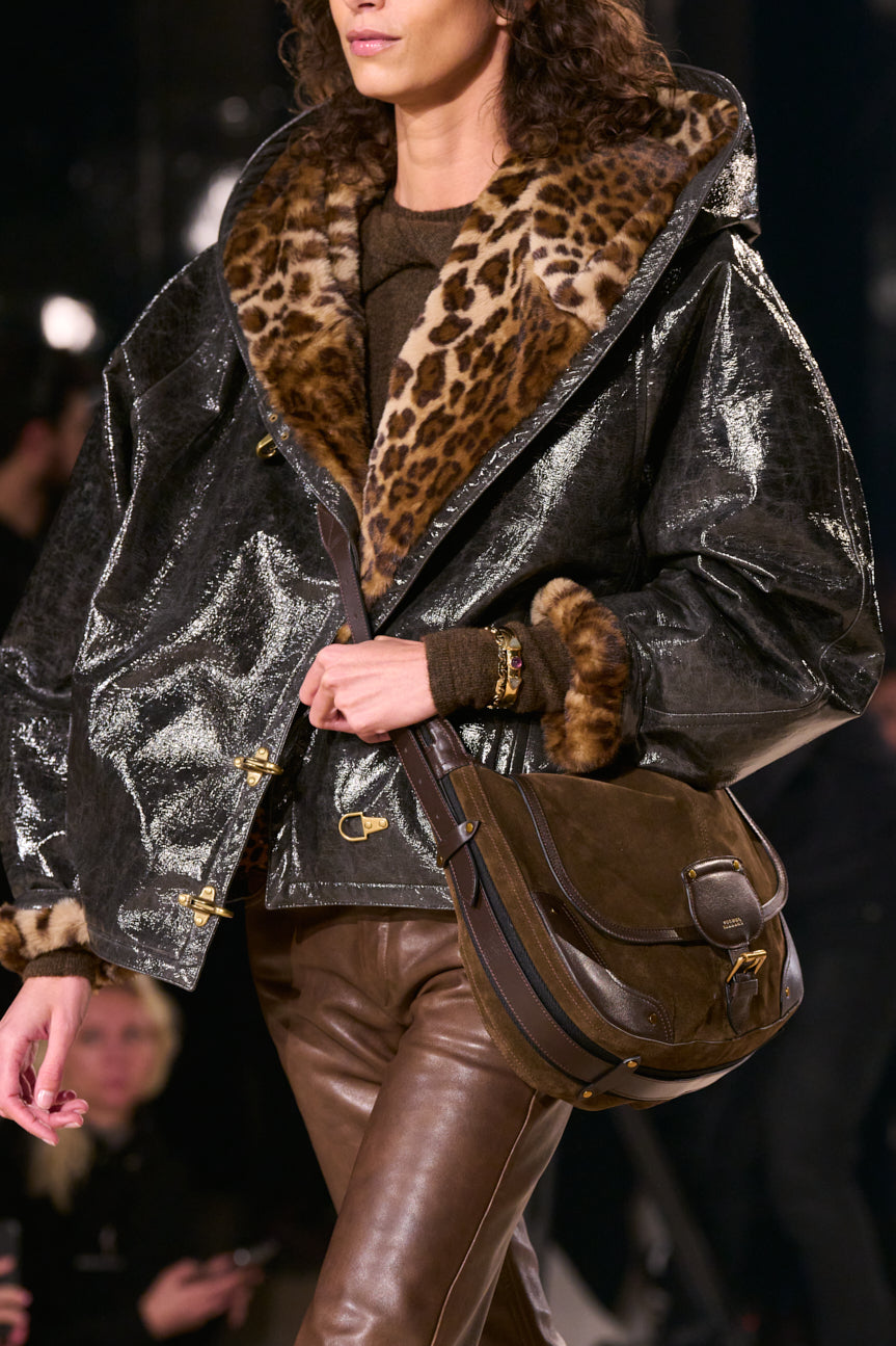 Cropped catwalk image of female model wearing a black coat lined in faux leopard and brown leather pants, carrying a brown messenger bag.