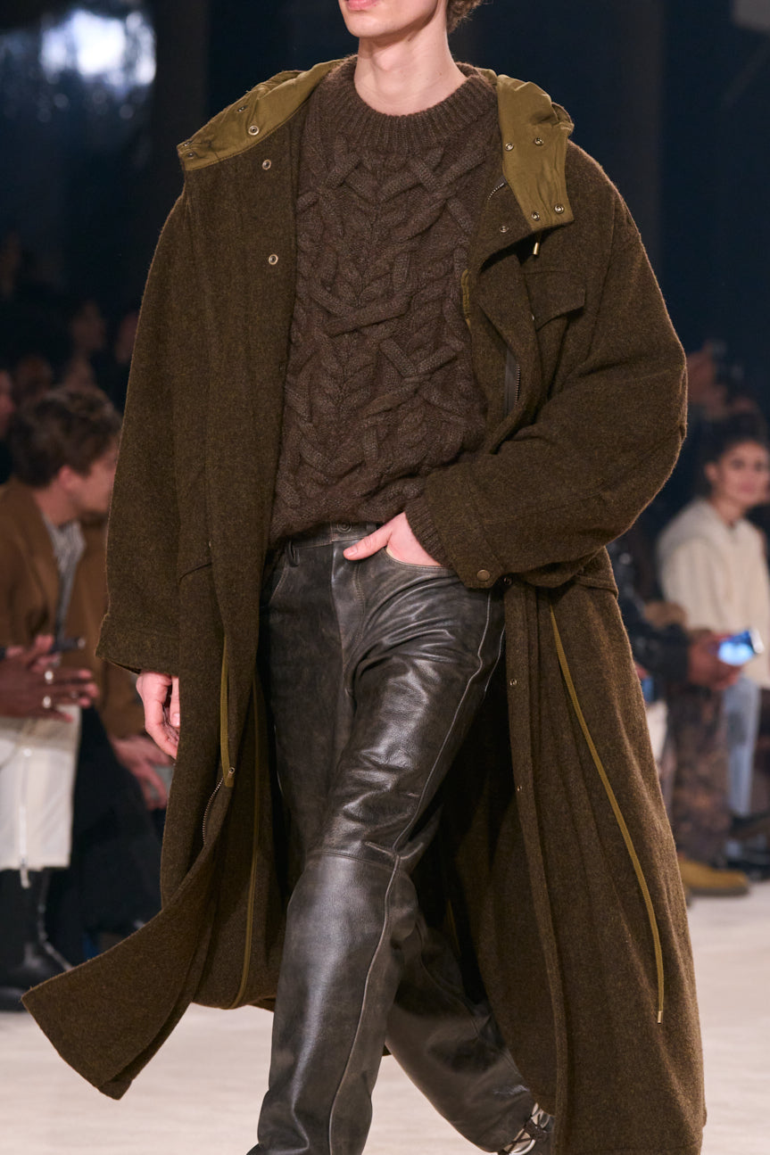 Cropped image of model on catwalk wearing a chunky brown cable-knit sweater, long chocolate brown coat and distressed brown leather jeans.