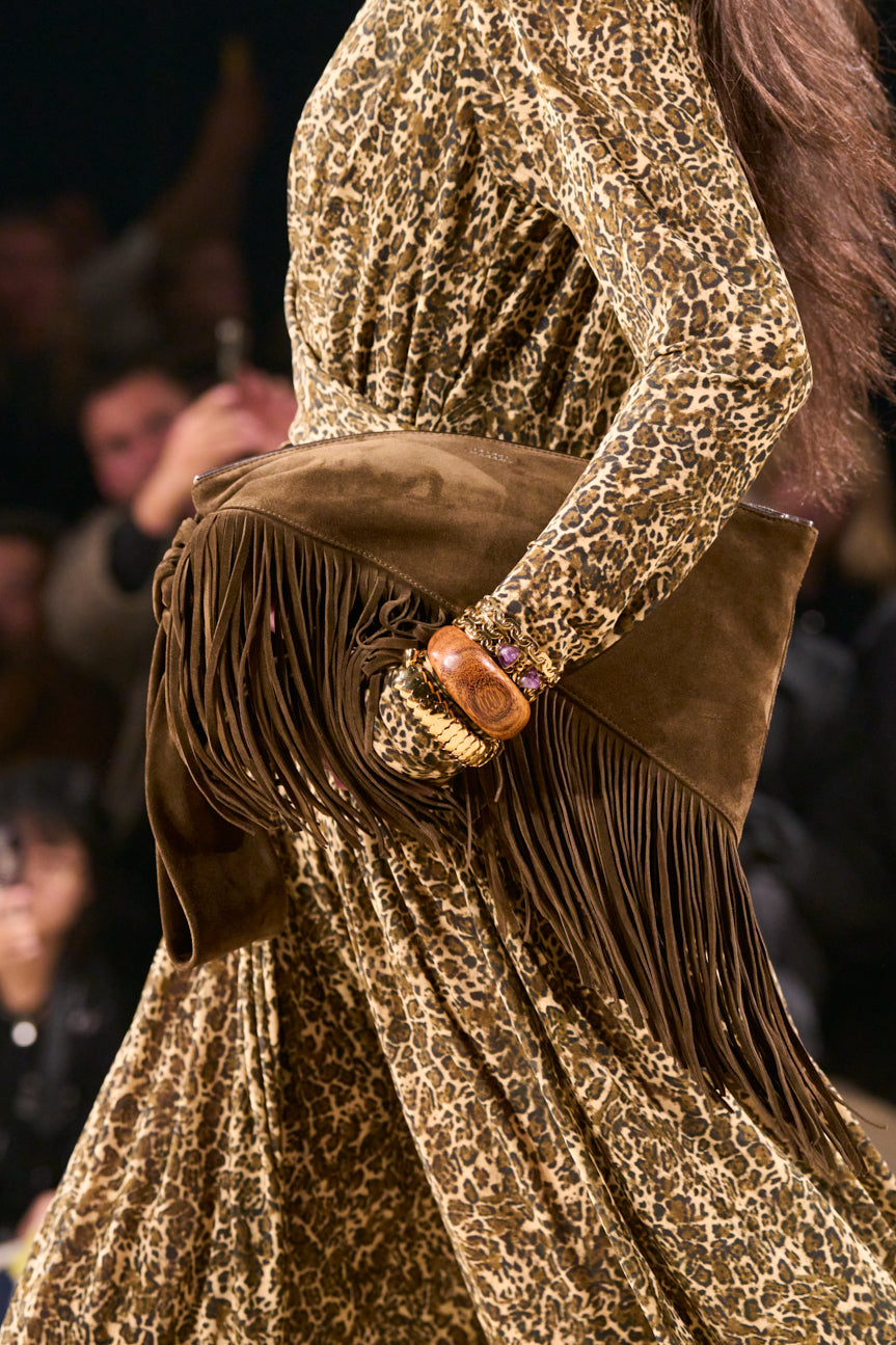 Cropped image of female midsection on catwalk wearing a leopard-printed gathered midi dress and carrying a large chocolate brown suede fringed bag.