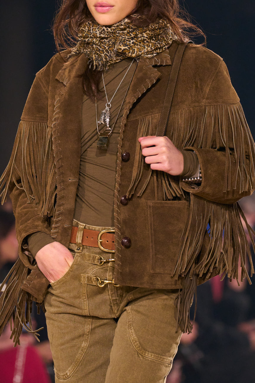 Cropped image of female model from chin to thigh, wearing a chocolate brown suede fringed jacket, leopard-printed scarf and washed khaki-colored jeans.