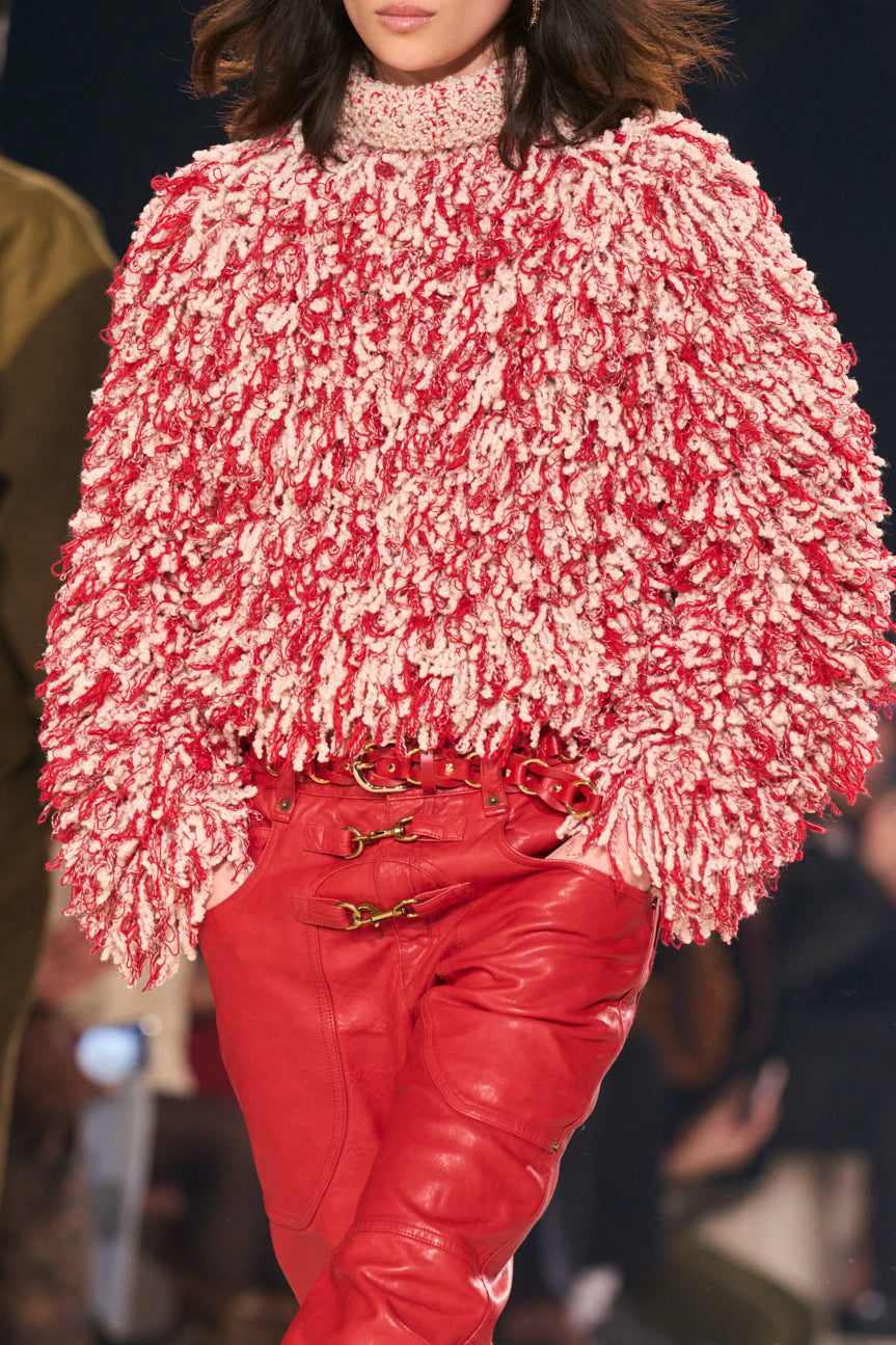 Cropped image from neck to thigh of female model on catwalk wearing a shaggy red and white oversized sweater and red leather pants.