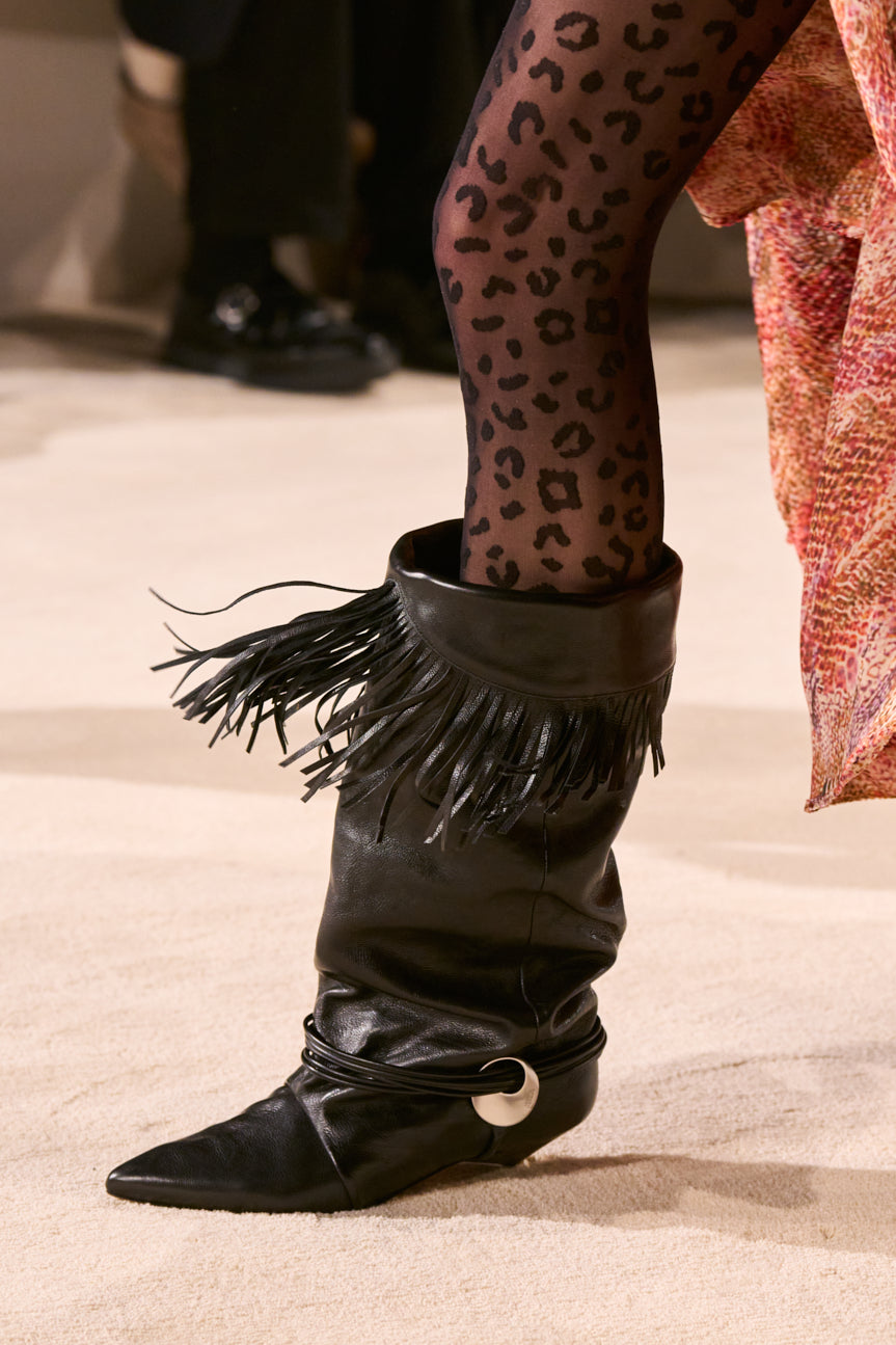 Cropped catwalk image of model’s leg in sheer black leopard-patterned tights and wearing a fringed black leather boot with a pointed toe.