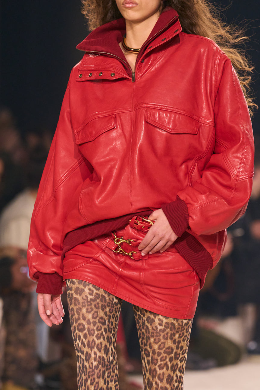 Cropped catwalk image of female model from chin to thigh wearing an oversized red leather top with matching miniskirt and leopard-printed tights.