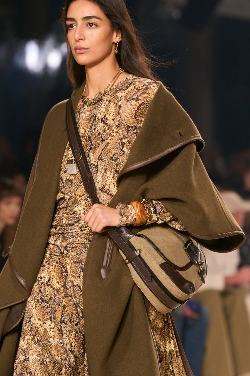 Cropped image of female model from head to mid-thigh wearing a tan snakeskin-printed midi dress, khaki-colored coat and carrying a tan messenger bag.
