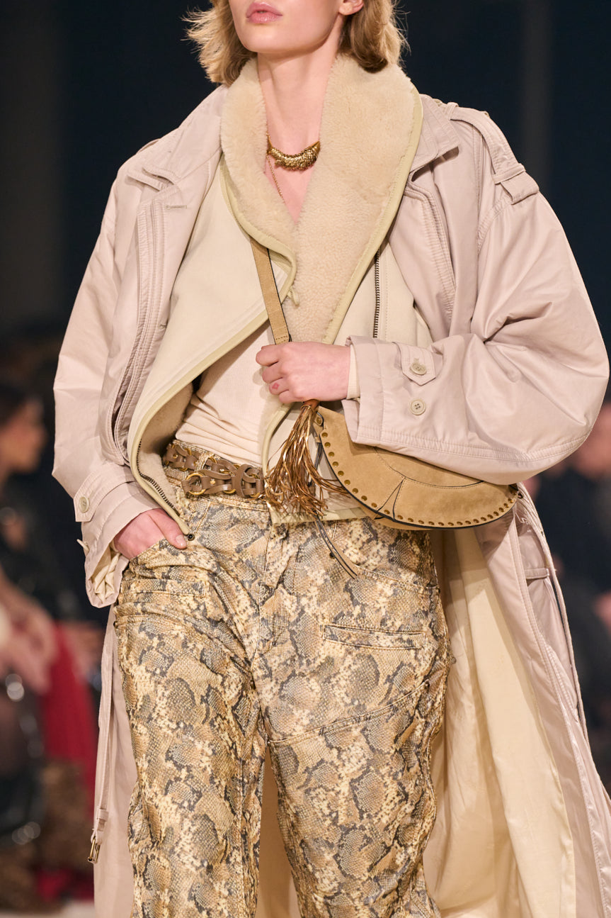 Cropped image of female model from chin to mid-thigh wearing an oversized blush-colored trench and tan snakeskin-printed pants with a brown braided belt.